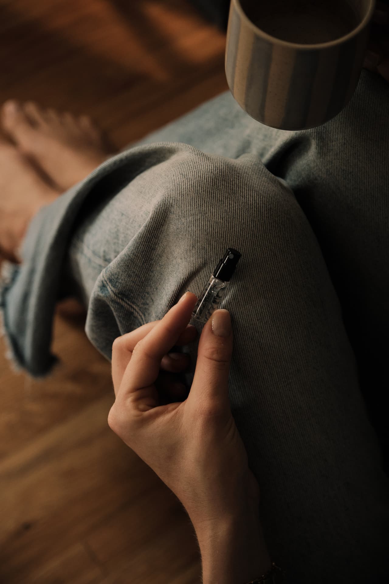 A hand holding a sample vial while sitting cross-legged in denim with a mug of coffee nearby