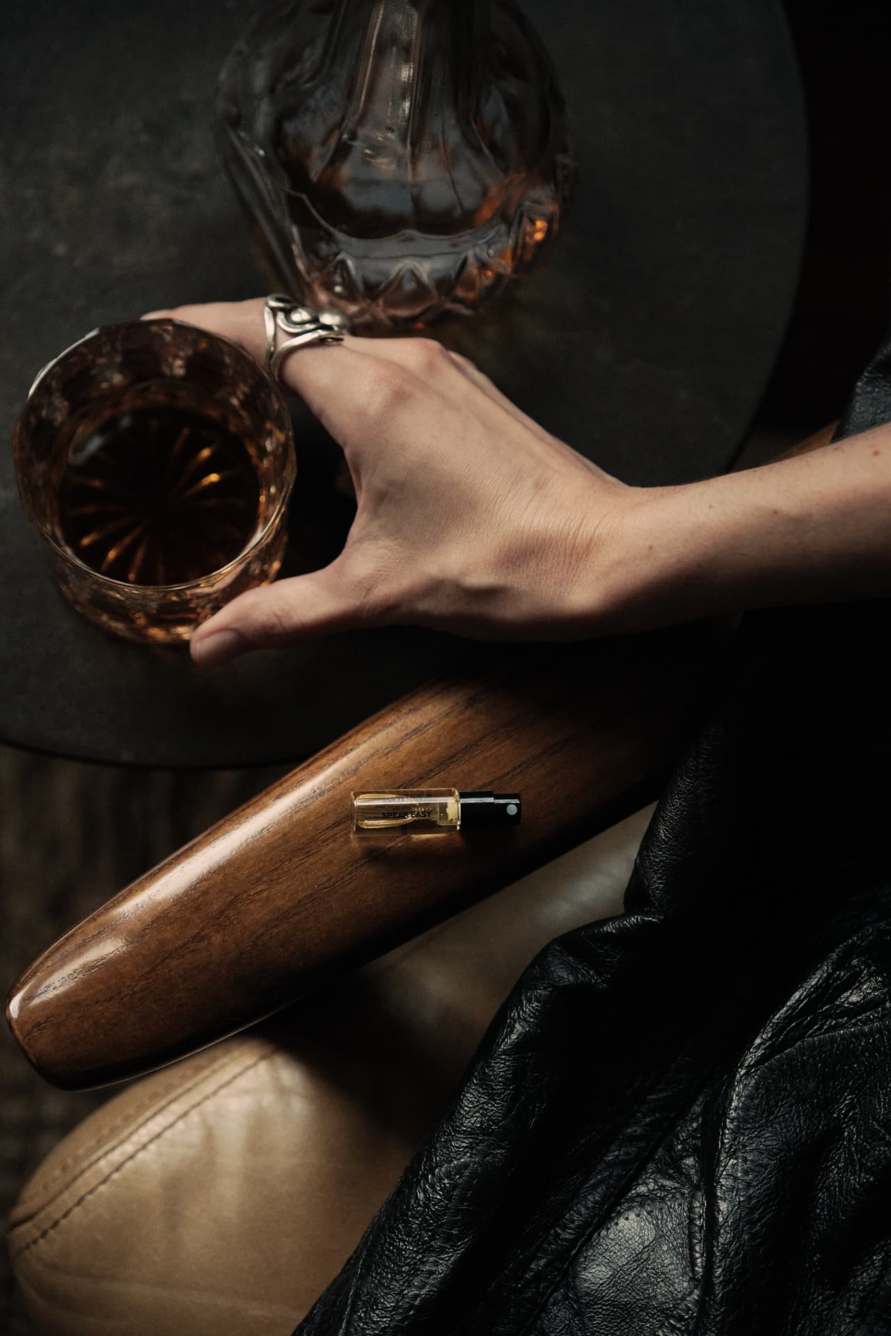 A Speakeasy sample vial resting on a wooden chair arm next to a glass of whiskey